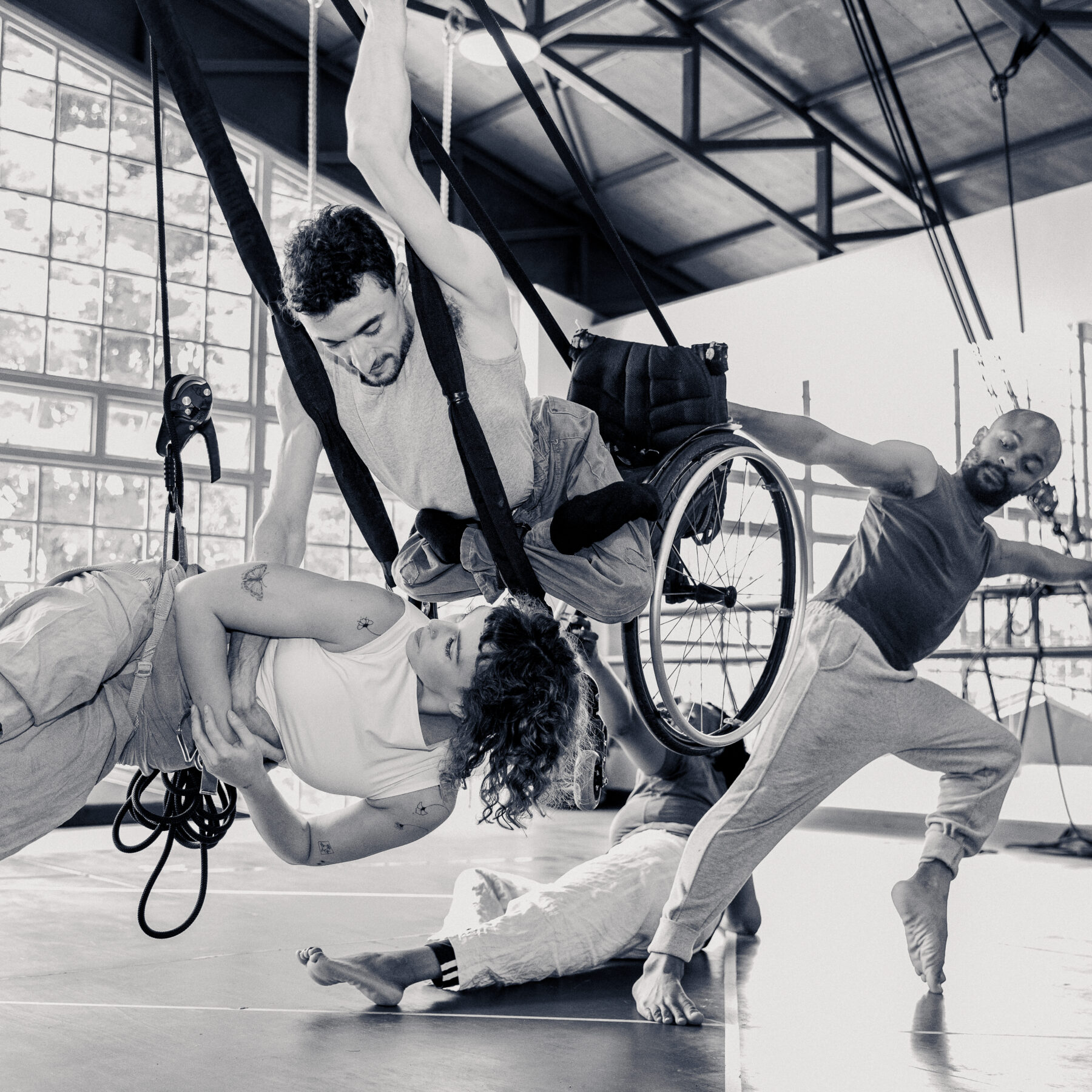 A group of diverse performers engage in an expressive aerial dance routine in a spacious studio with large windows. One performer, suspended in a wheelchair with the support of harnesses, gracefully lifts a fellow dancer who reaches towards him. Another dancer balances on one foot with an outstretched arm, adding to the dynamic movement. The composition captures strength, collaboration, and inclusivity in an artistic performance.