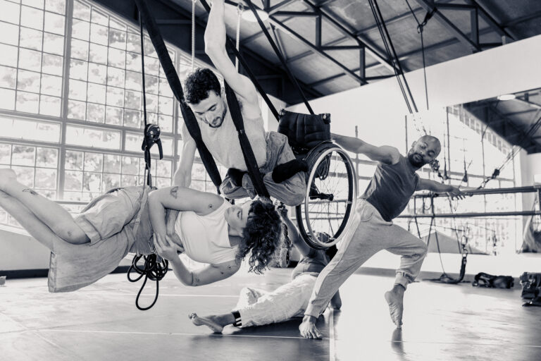 A group of diverse performers engage in an expressive aerial dance routine in a spacious studio with large windows. One performer, suspended in a wheelchair with the support of harnesses, gracefully lifts a fellow dancer who reaches towards him. Another dancer balances on one foot with an outstretched arm, adding to the dynamic movement. The composition captures strength, collaboration, and inclusivity in an artistic performance.