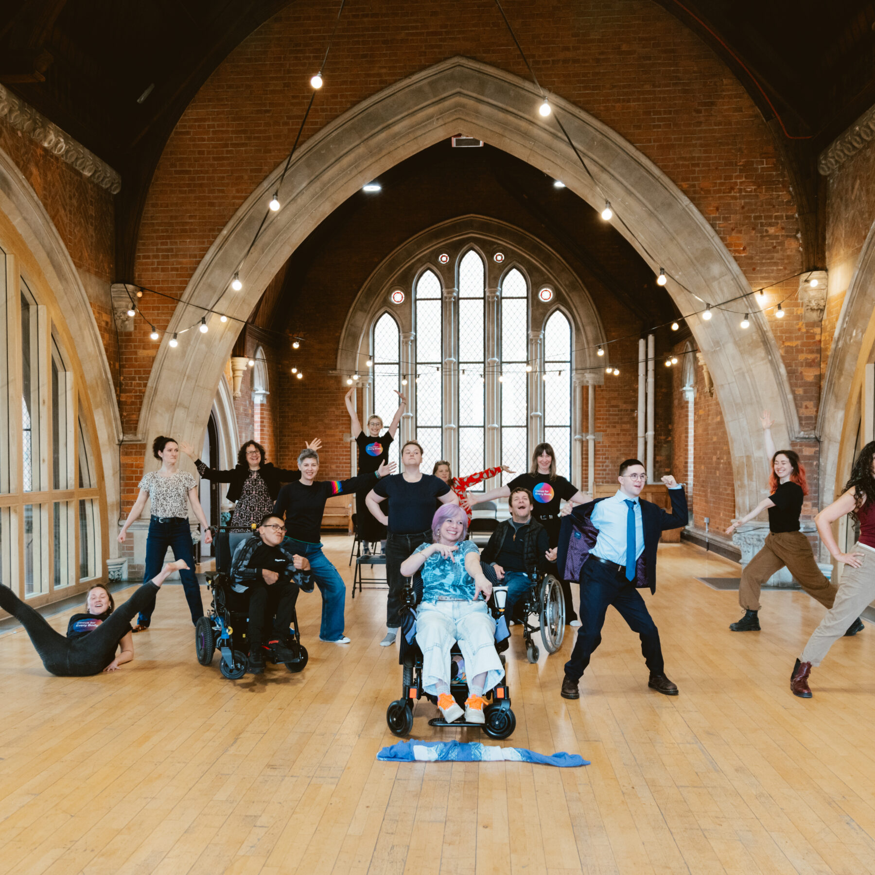 A group of performers pose for a photo in a beautiful rehearsal space with arched windows. Connecting Communities